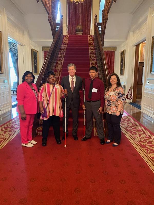 Two GMS students holding their white canes stand with NC Governor Roy Cooper in front of the main staircase in the Governor's Mansion. Included in the picture are the adult sponsors, Dr. McManus and Ms. Medina.